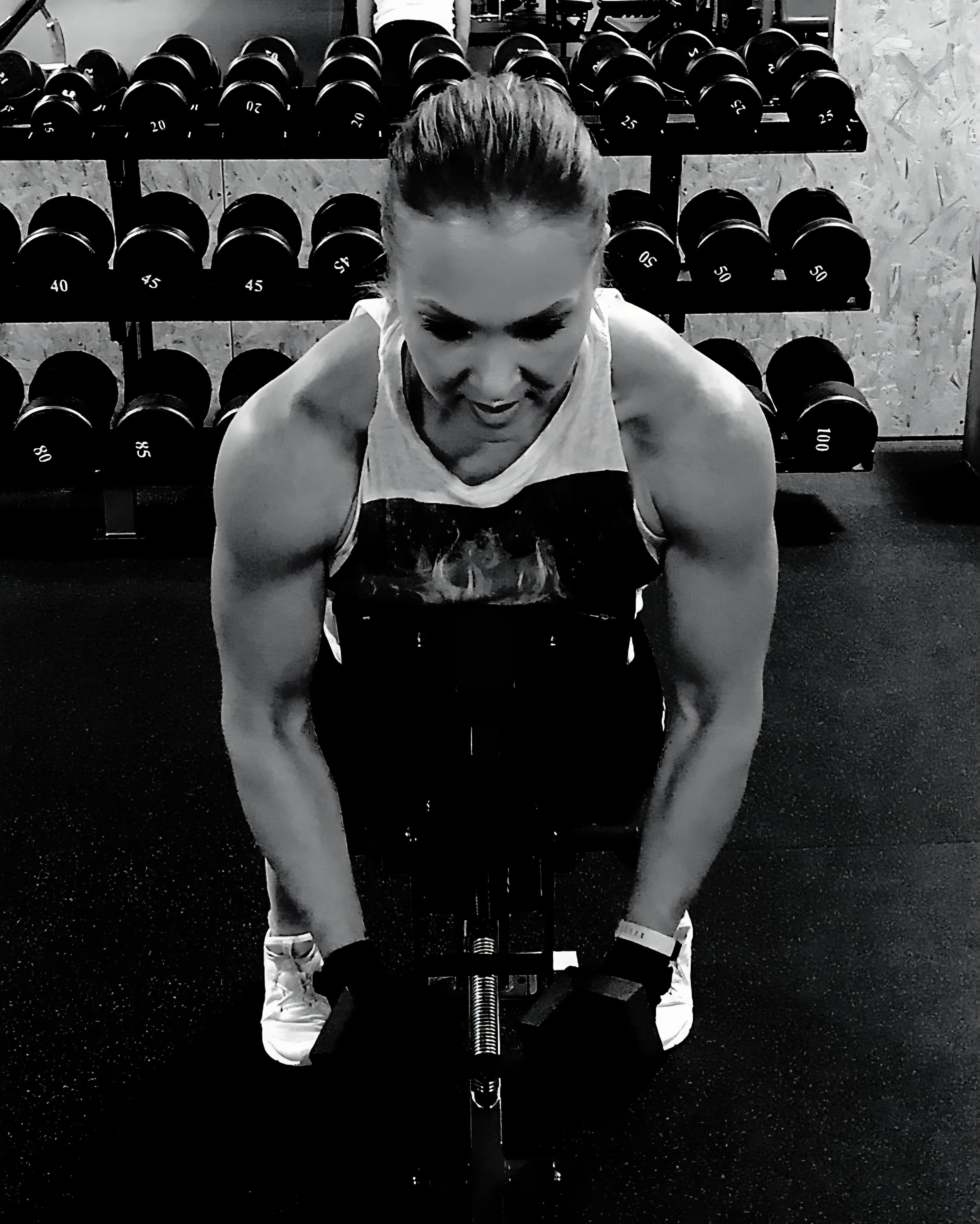 Muscular woman in black and white, focused on gym exercise with dumbbells in background.
