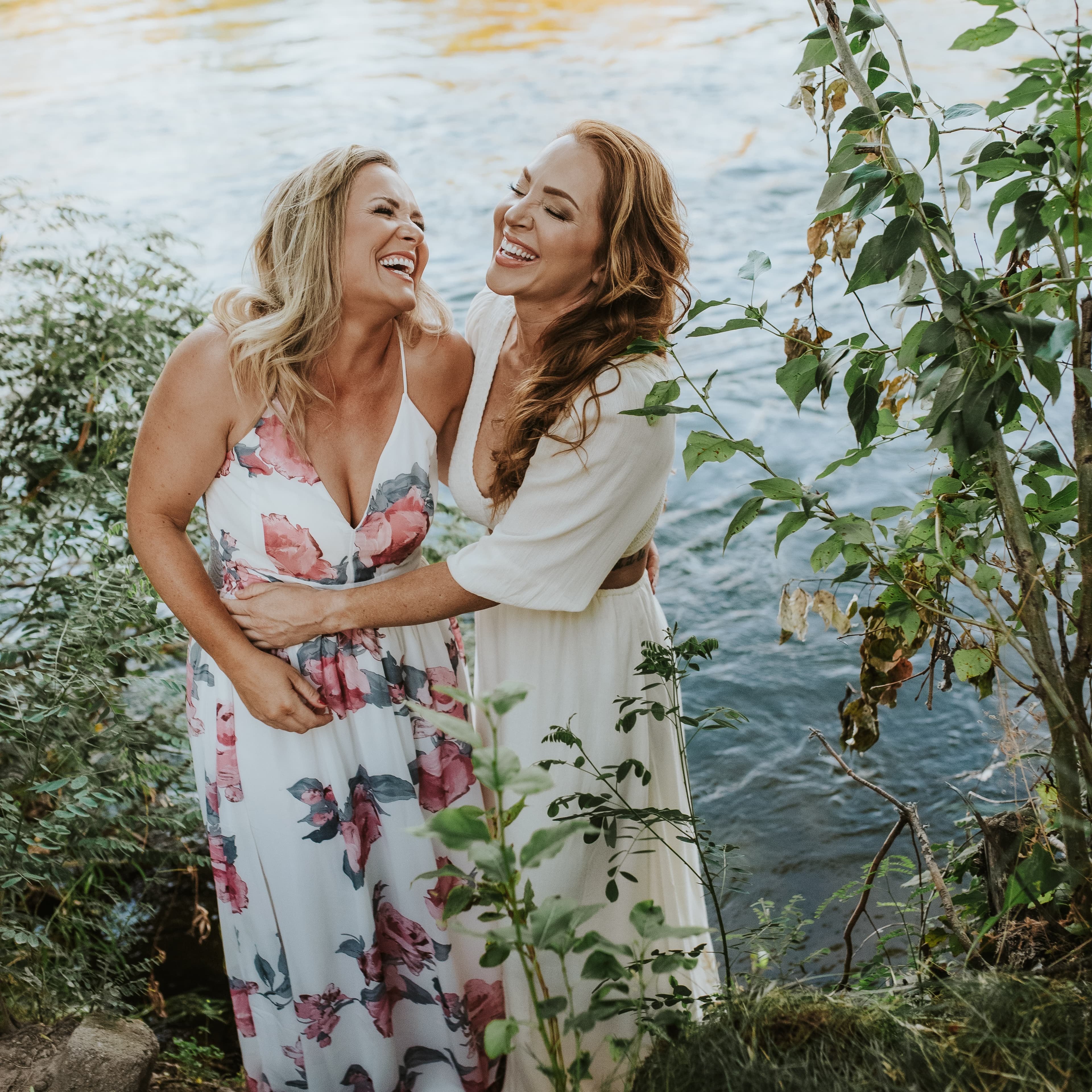 Two women laughing joyfully, embracing by a riverbank amidst green foliage.